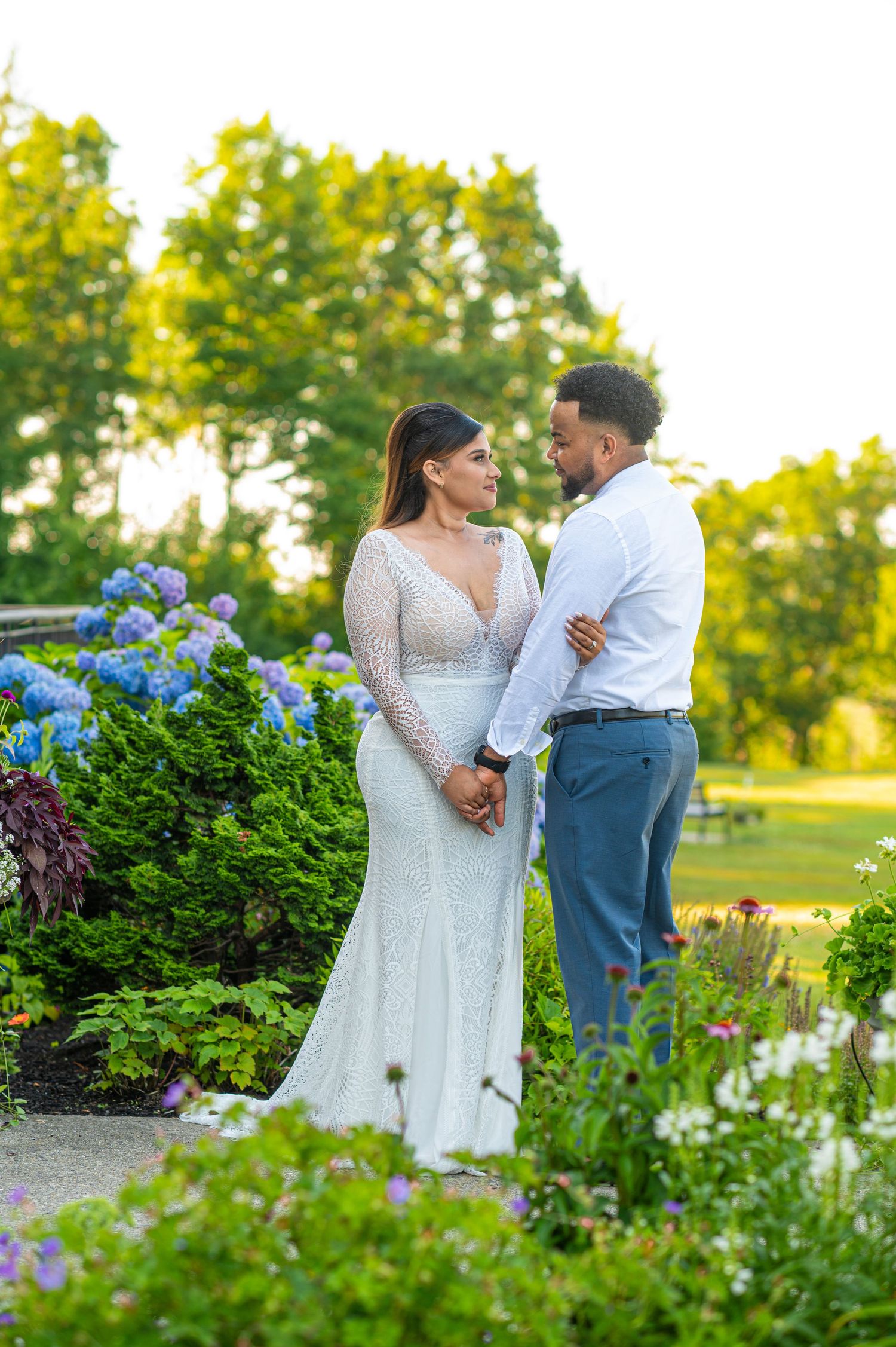 Couple in wedding attire holding hands in a garden with blue hydrangeas and green trees at golden hour.
