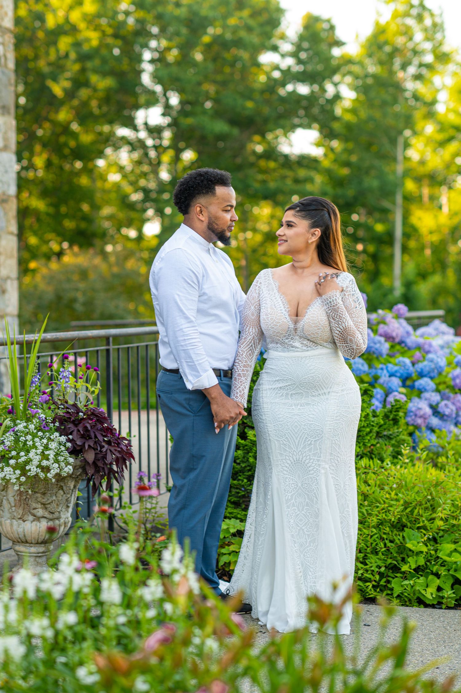 Couple in wedding attire holding hands in a lush garden with blue hydrangeas blooming in the background.