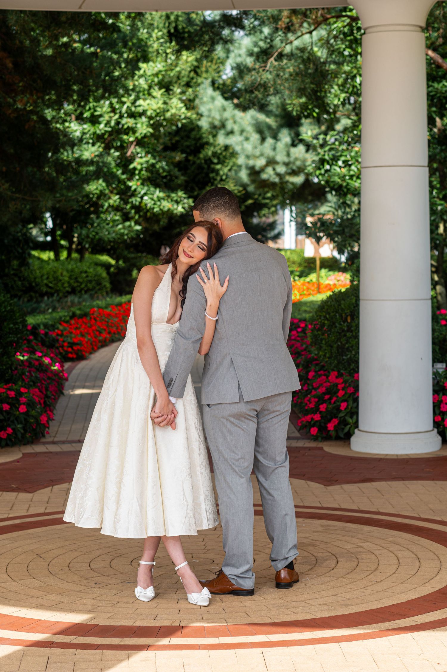 Bride in white tea-length dress and groom in gray suit share a romantic embrace under a garden pavilion with colorful flowers.