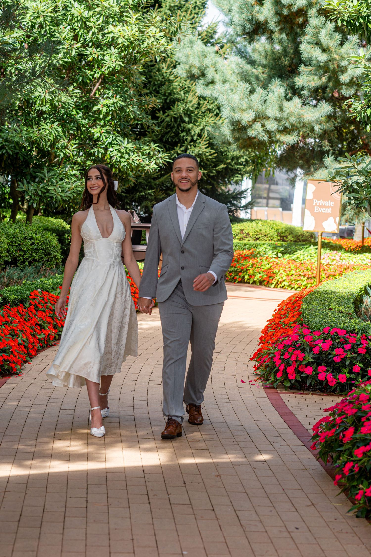 Couple holding hands walking on brick path through colorful flower gardens, woman in white dress, man in gray suit.