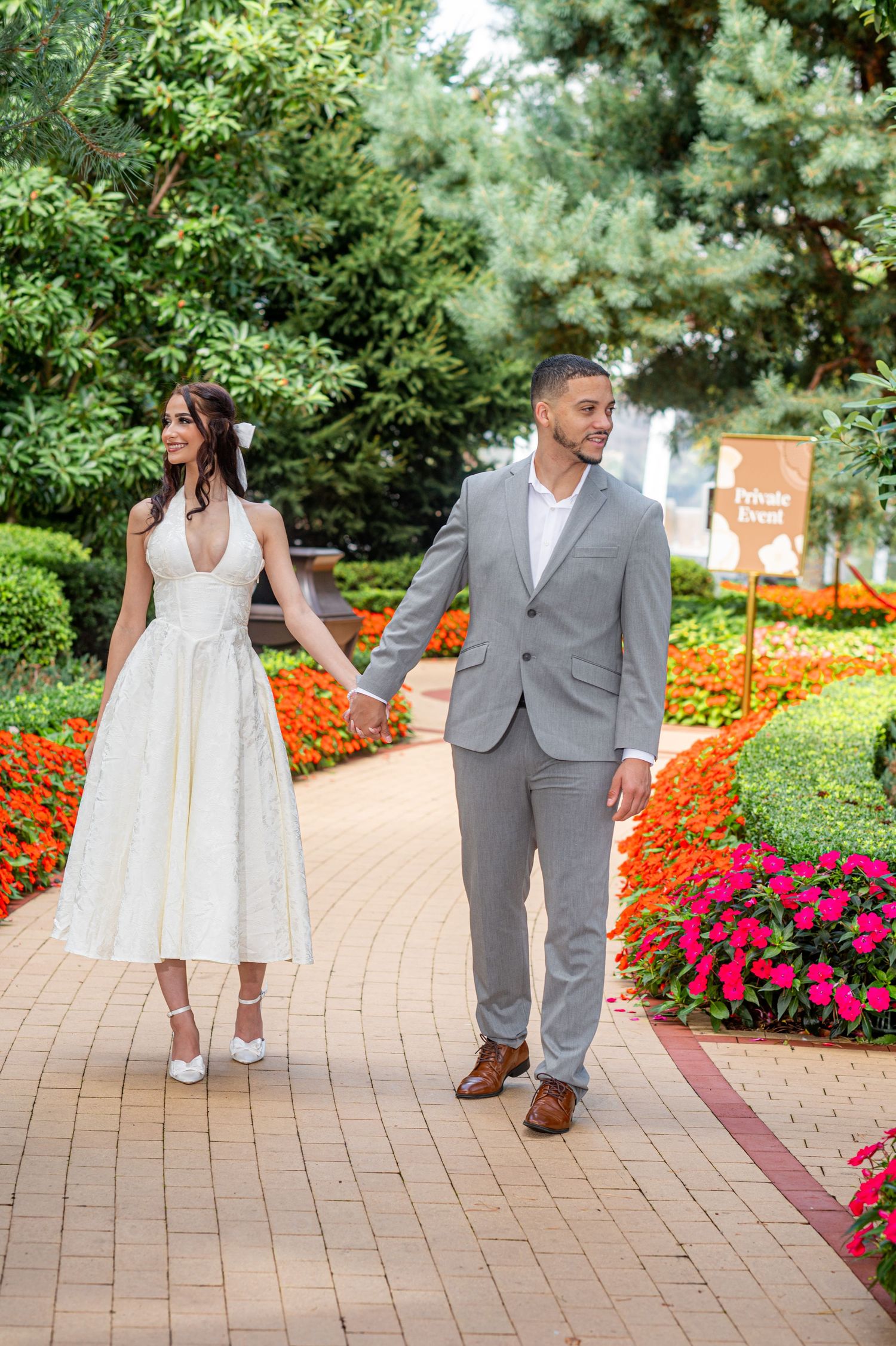 Couple in wedding attire holding hands walking through a colorful garden path with blooming flowers.