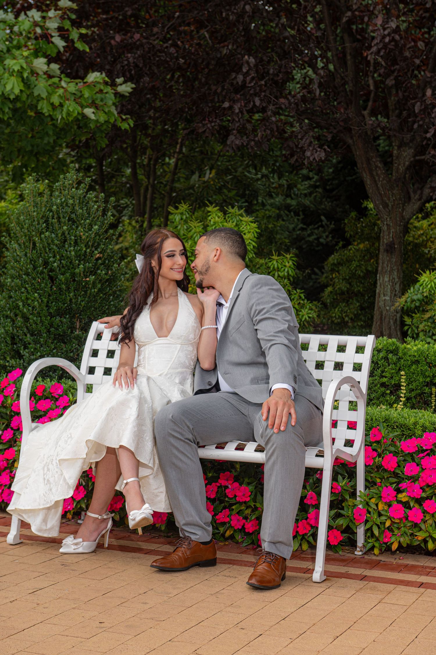 Bride and groom sitting on white bench surrounded by pink flowers in a lush garden setting.