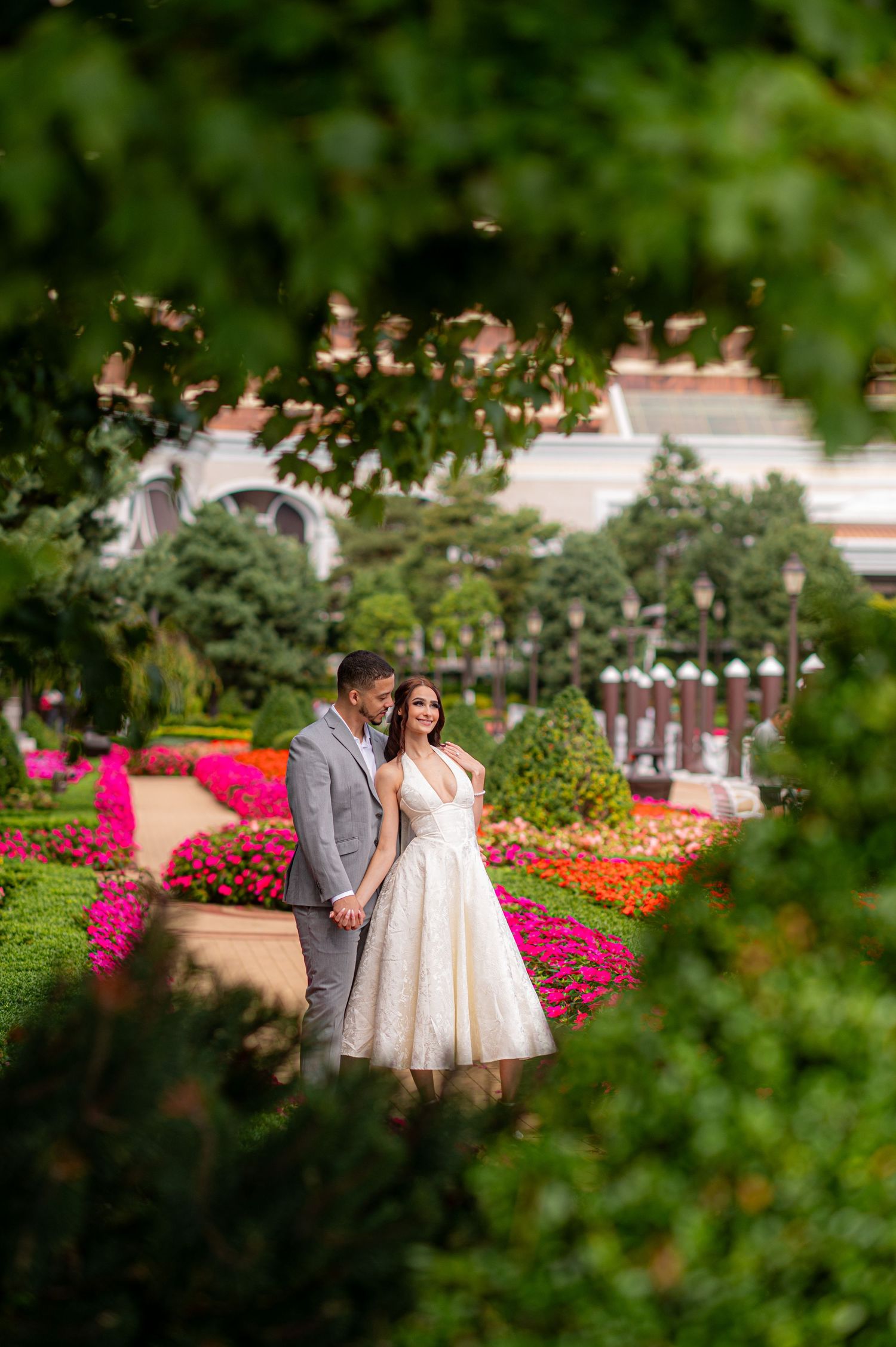 Couple in wedding attire posing in a lush garden with colorful flowers and green trees framing the romantic scene.