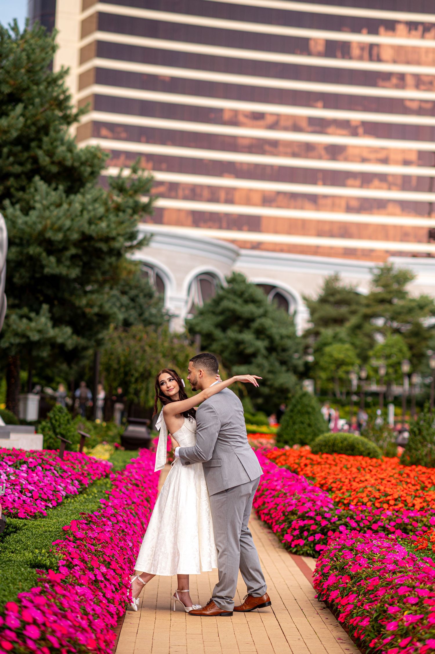 Couple dancing on a flower-lined path with vibrant blooms and a grand building in background.