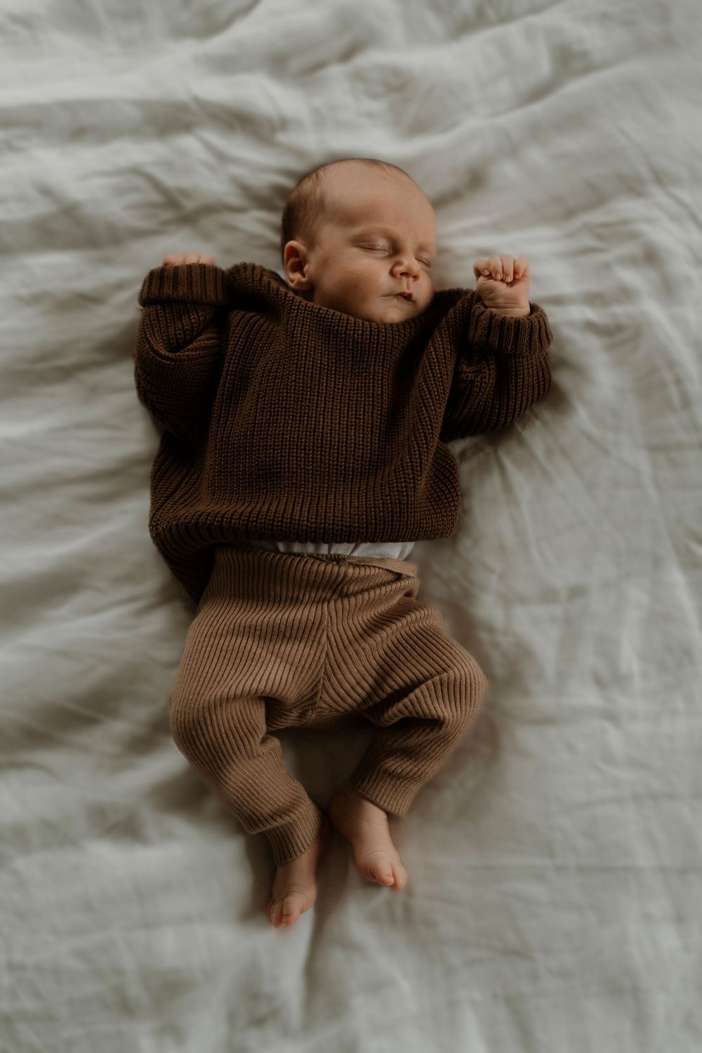 A newborn baby wearing brown knit clothing lies peacefully on soft white bedding.