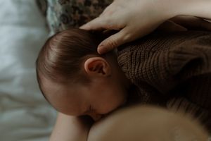 A baby in a brown knit sweater naps peacefully on soft white bedding.