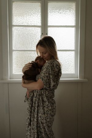 Figure in a floral dress stands by a frosted window holding a small pet in a tender embrace.