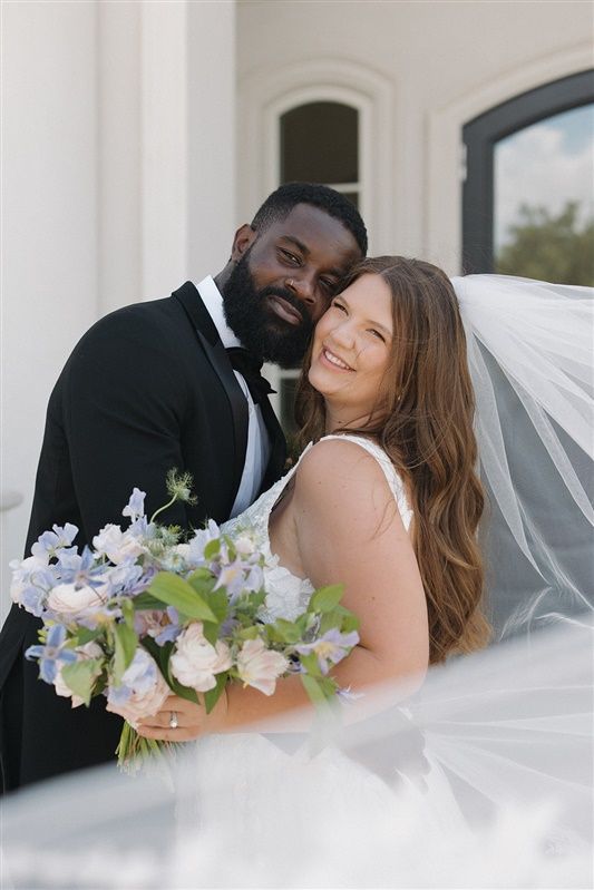 A wedding couple sharing an intimate moment with a white veil and light purple bouquet in front of a building.