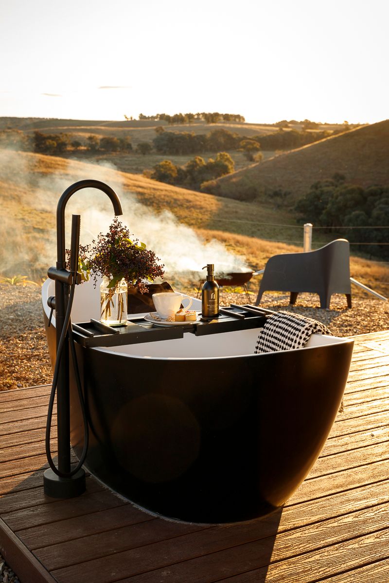Luxury tiny house with an outdoor bath and fire pit looking out to a view of rolling hills at sunset. Property photography by Em Callaghan.
