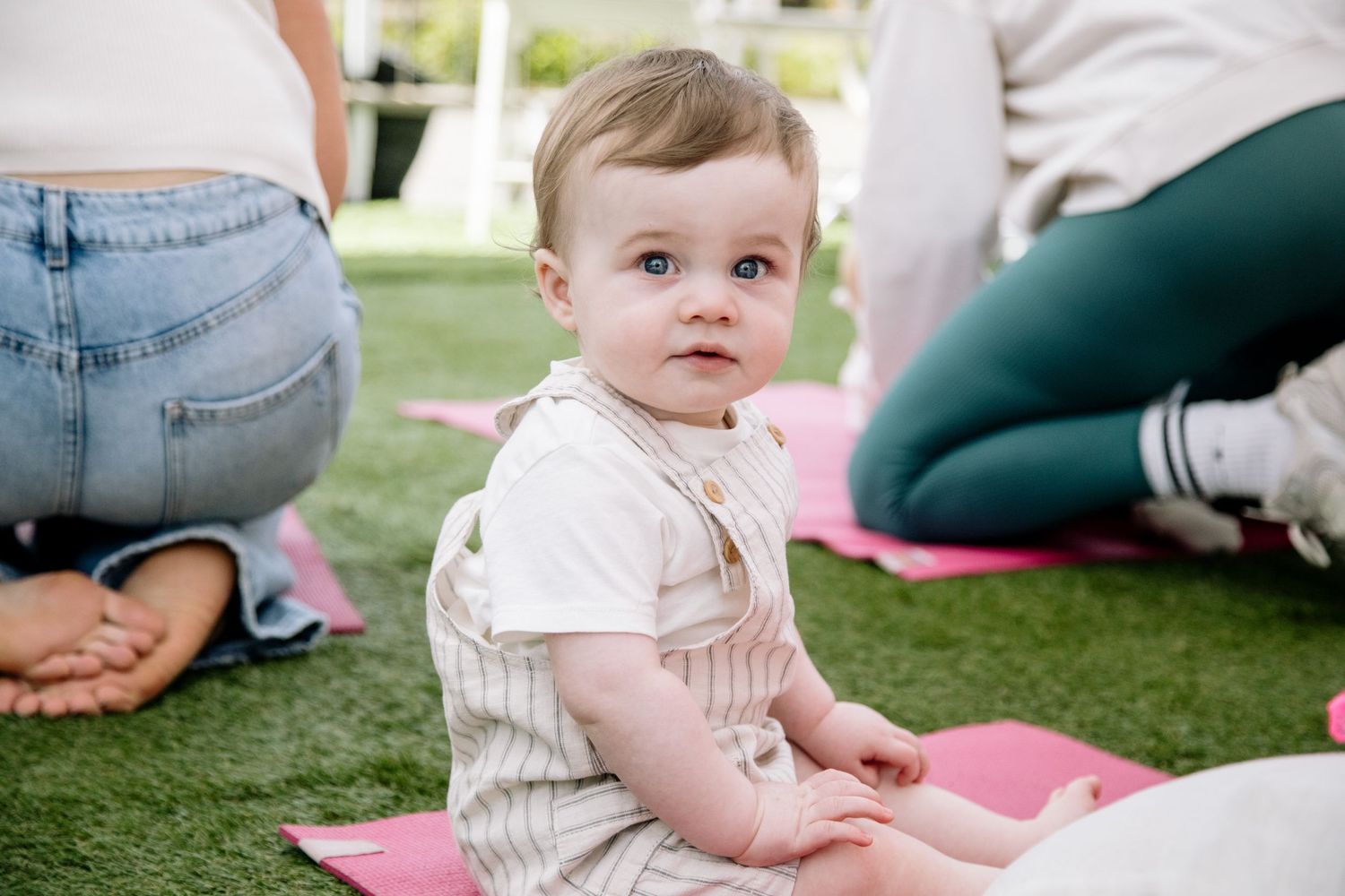 Baby in light colored clothing sits attentively on a pink exercise mat during an outdoor class.