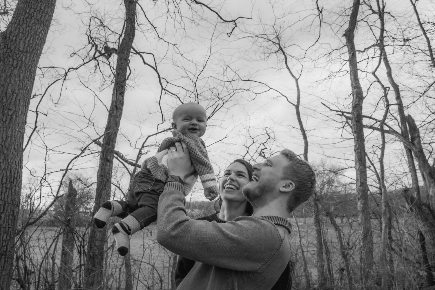 An outdoor winter scene shows a joyful moment with a small child being lifted up against a background of bare trees.