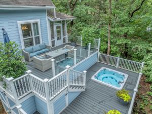Multi-level deck with hot tub and seating areas attached to blue house overlooking wooded area.