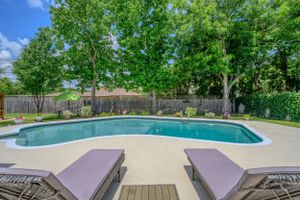Curved swimming pool with concrete deck and lounge chairs surrounded by lush green trees.