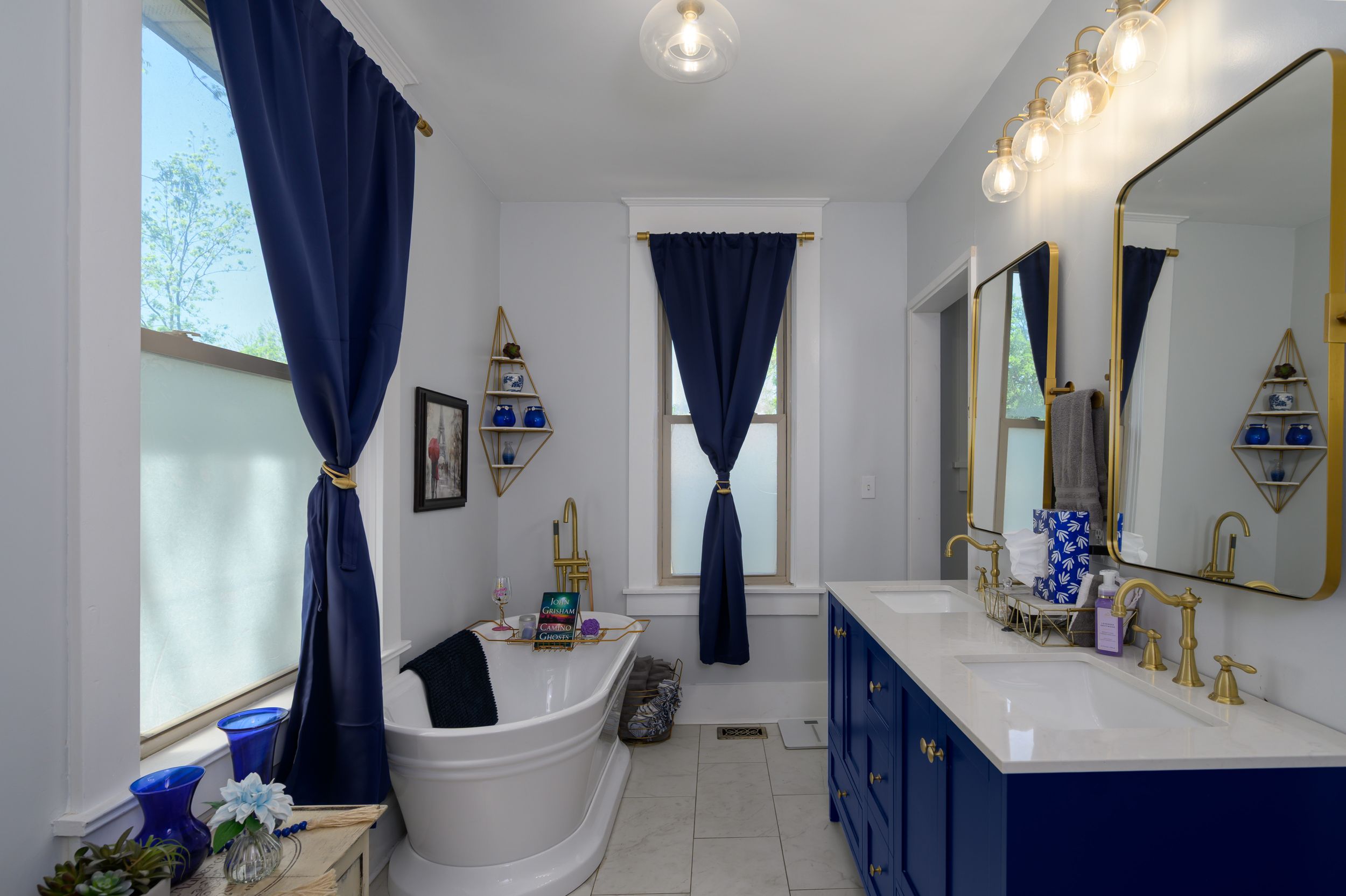 A bright modern bathroom design features navy blue accents, gold fixtures, and a freestanding white bathtub near windows.