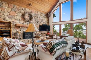 Rustic mountain lodge living room with stone walls, vaulted wood ceiling, and panoramic windows overlooking scenic views.