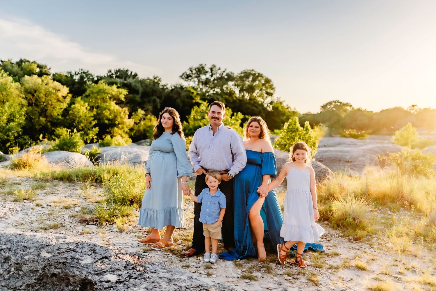 Family in coordinated blue and white outfits pose together at sunset in a natural outdoor setting.
