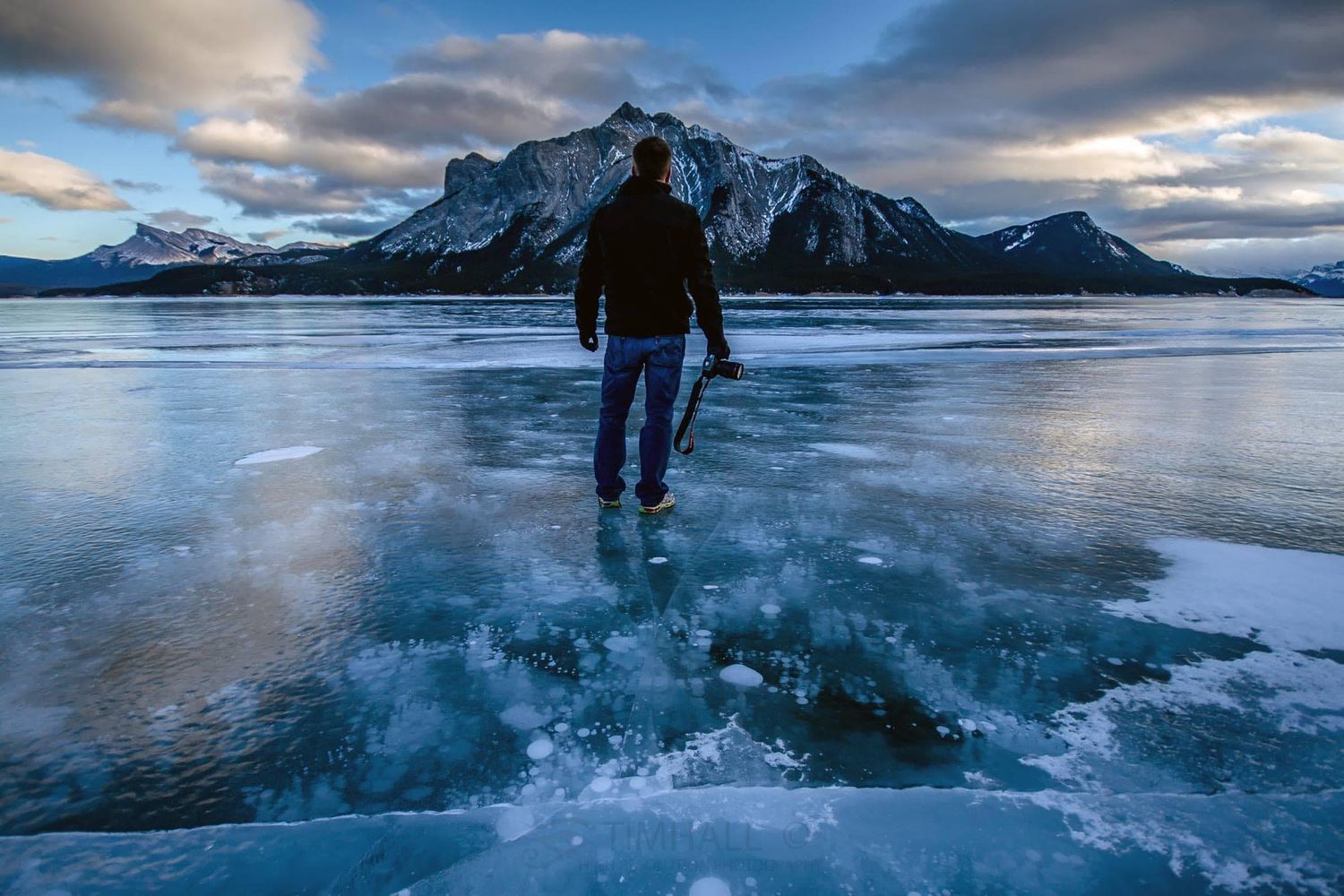 Man standing on frozen lake with snow-capped mountains in background at dusk in Banff, Alberta.