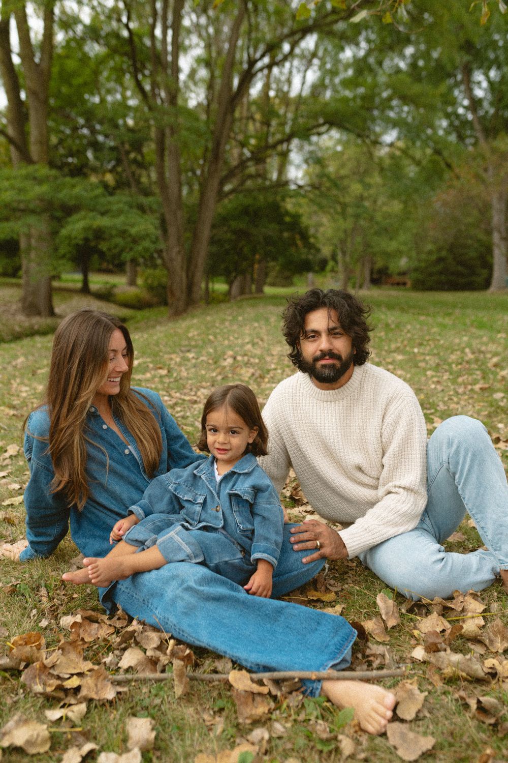 Family lying together in autumn leaves wearing denim and cream sweater in outdoor park setting.