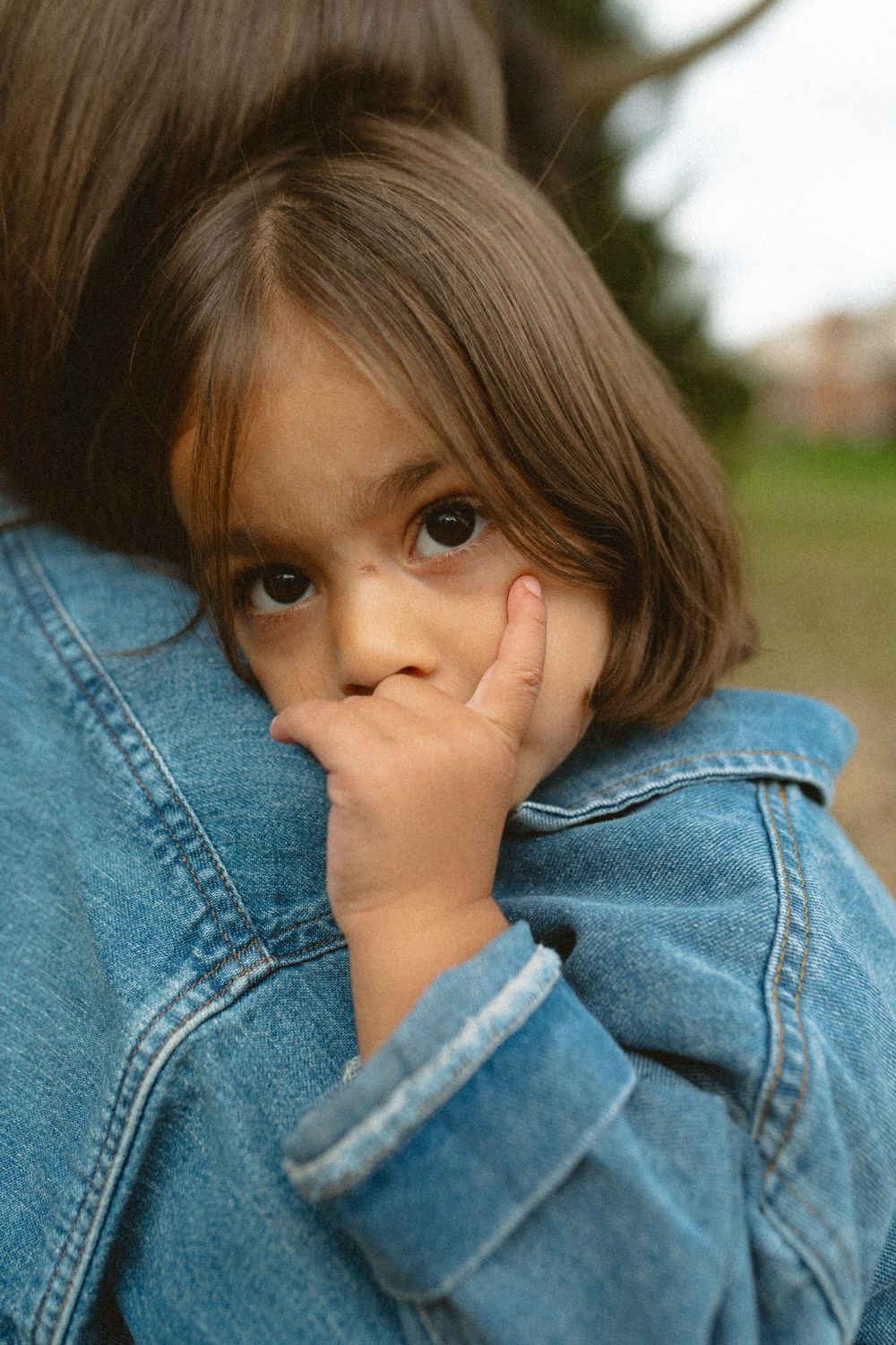 A child in a denim jacket peeks shyly at the camera while covering their face with their sleeve.