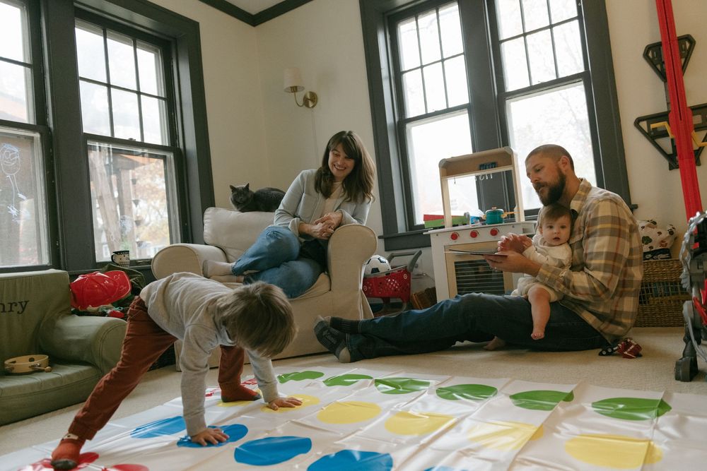 Family plays energetic game of Twister together on colorful mat in living room with large windows.
