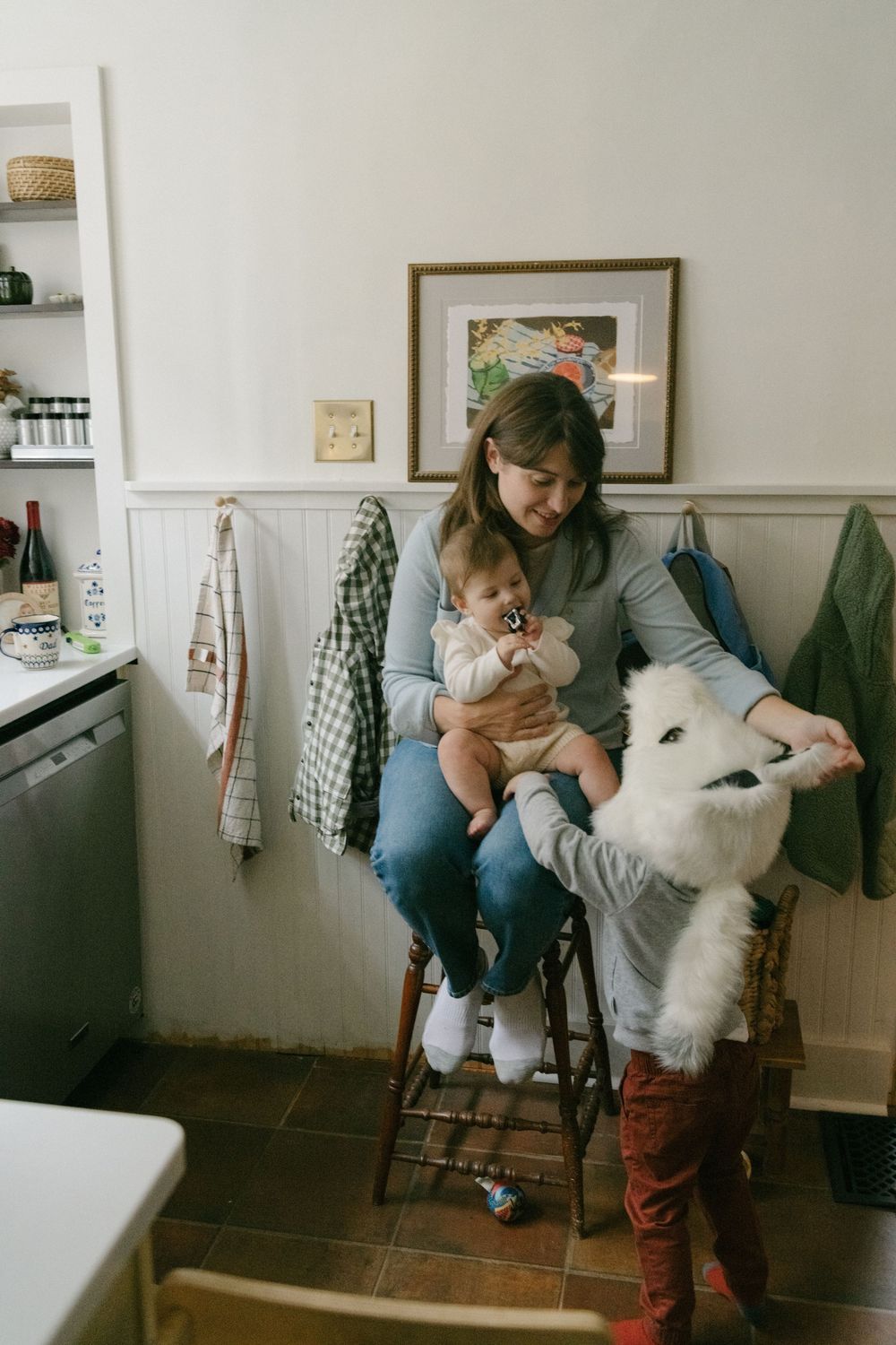 A cozy kitchen scene shows a person sitting on a stool with a baby and a large white dog beside them.