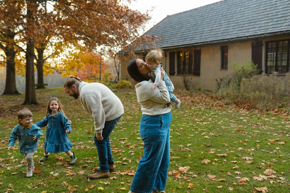People in teal pants gather fallen autumn leaves on a residential lawn during a fall cleanup.