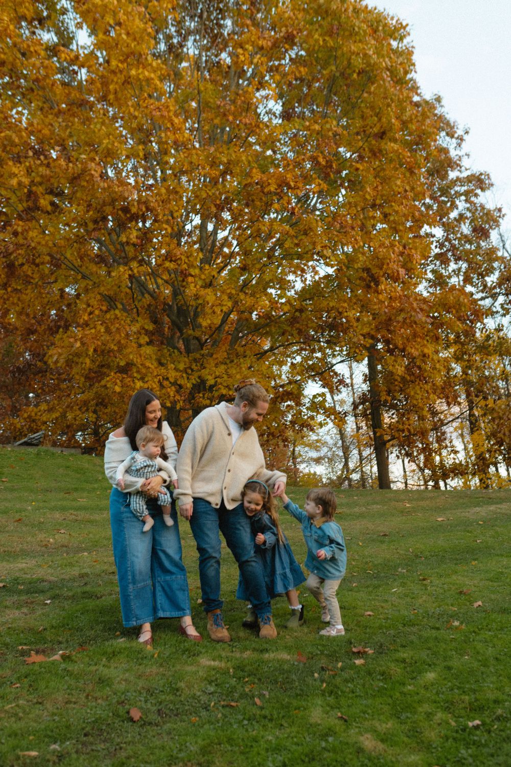 Family wearing coordinating blue outfits sits together on grassy hill during golden autumn sunset.