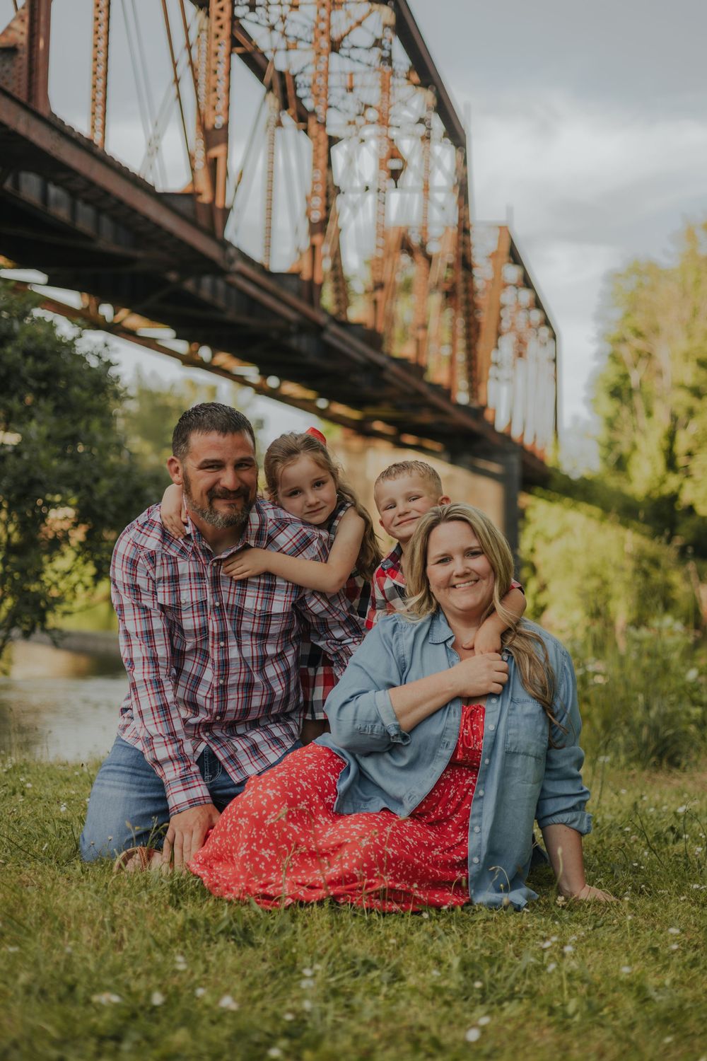 A family portrait taken beside an old iron bridge on a grassy riverbank.
