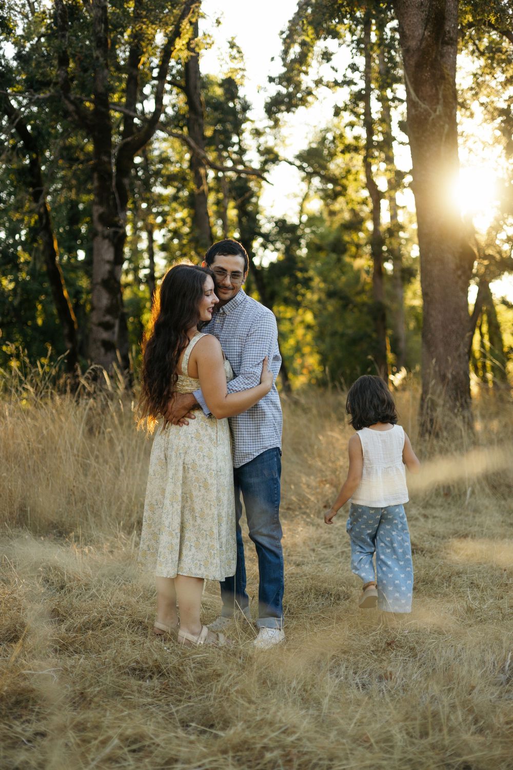 A picturesque family portrait session in a golden field at sunset with tall trees in the background.