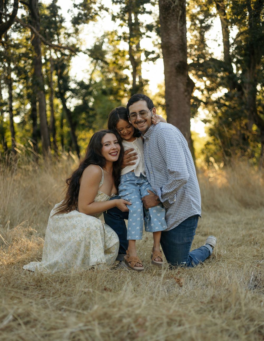 A loving family shares candid moments in a sunlit meadow at golden hour amid tall grass and trees.