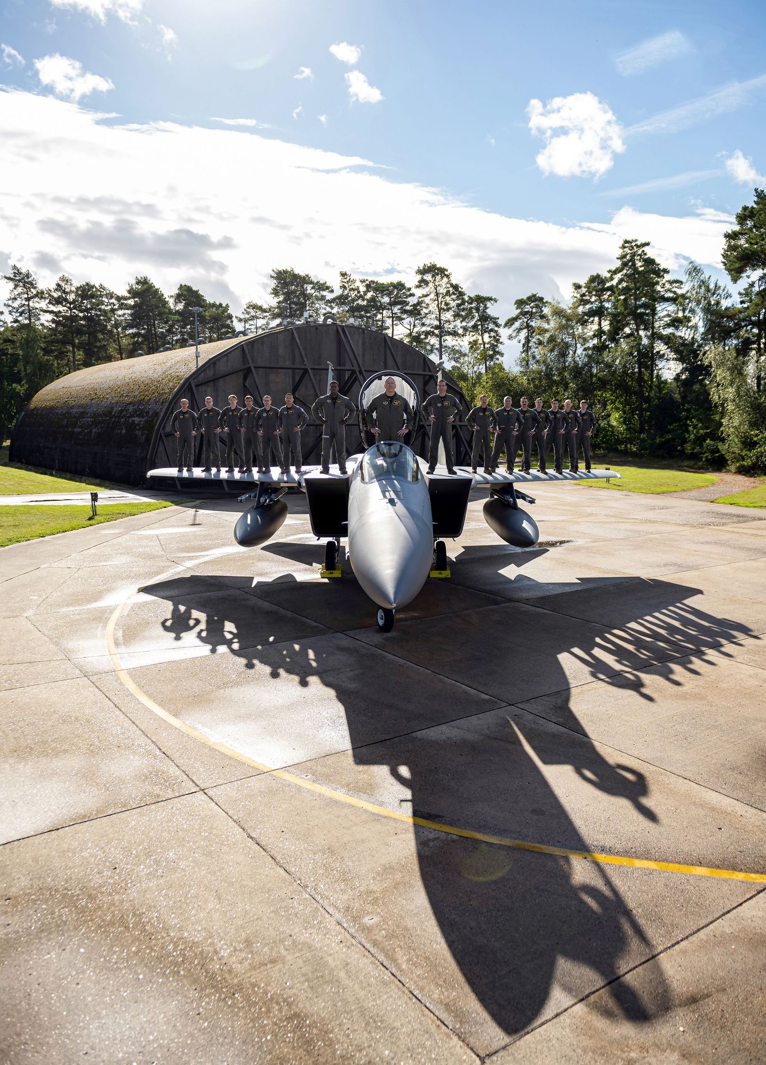 Military jet fighter parked on tarmac with uniformed personnel lined up in front of a camouflaged hangar surrounded by trees.