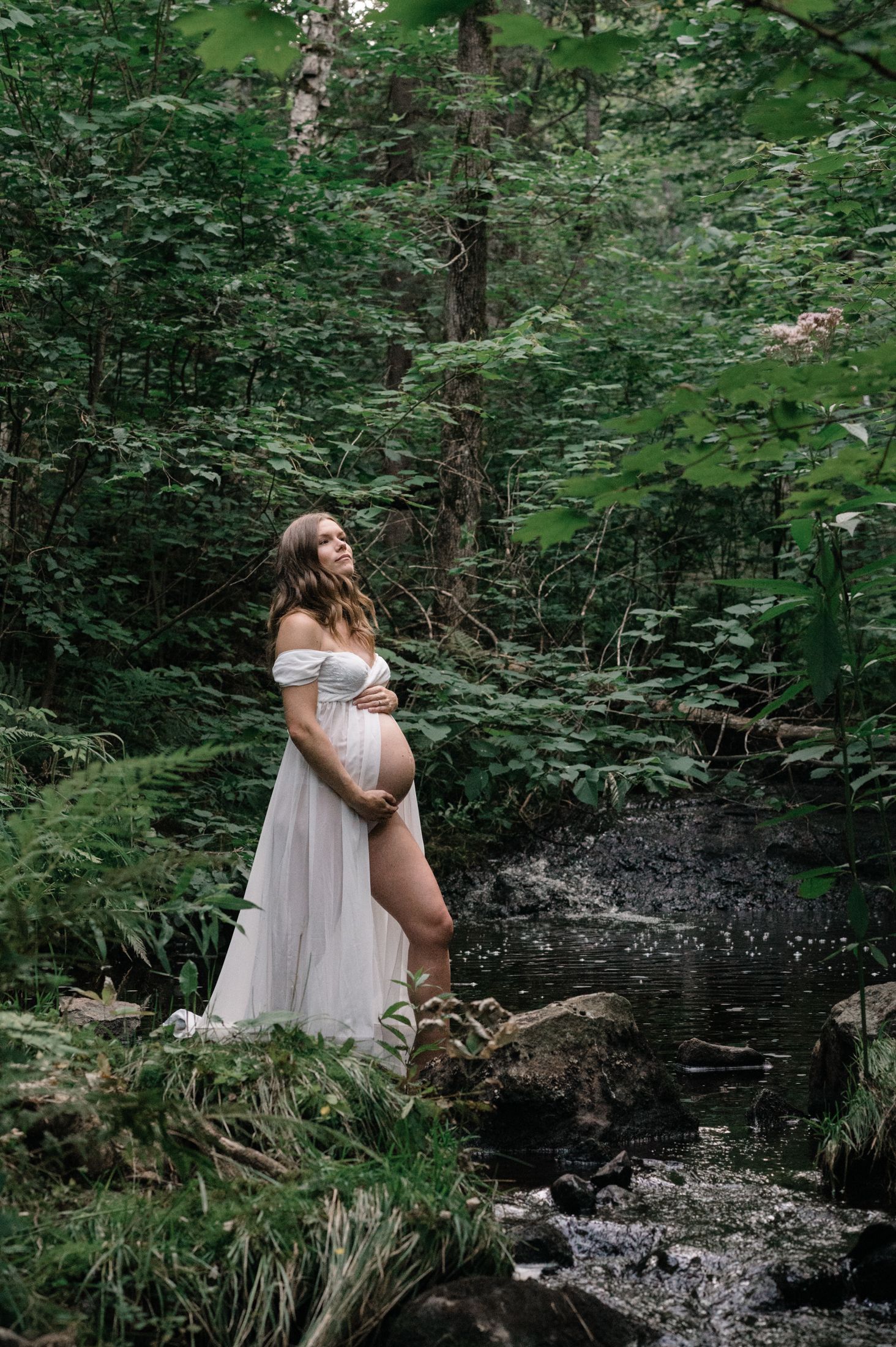 Maternity photo of woman in white dress standing by forest stream.