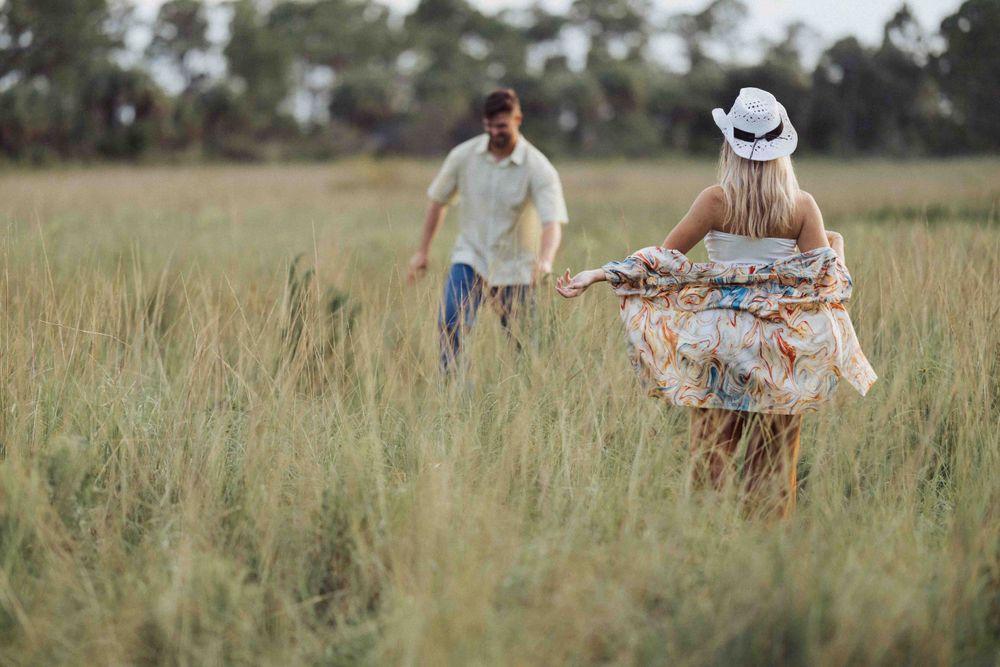 A couple plays chase through a tall grass field at sunset.