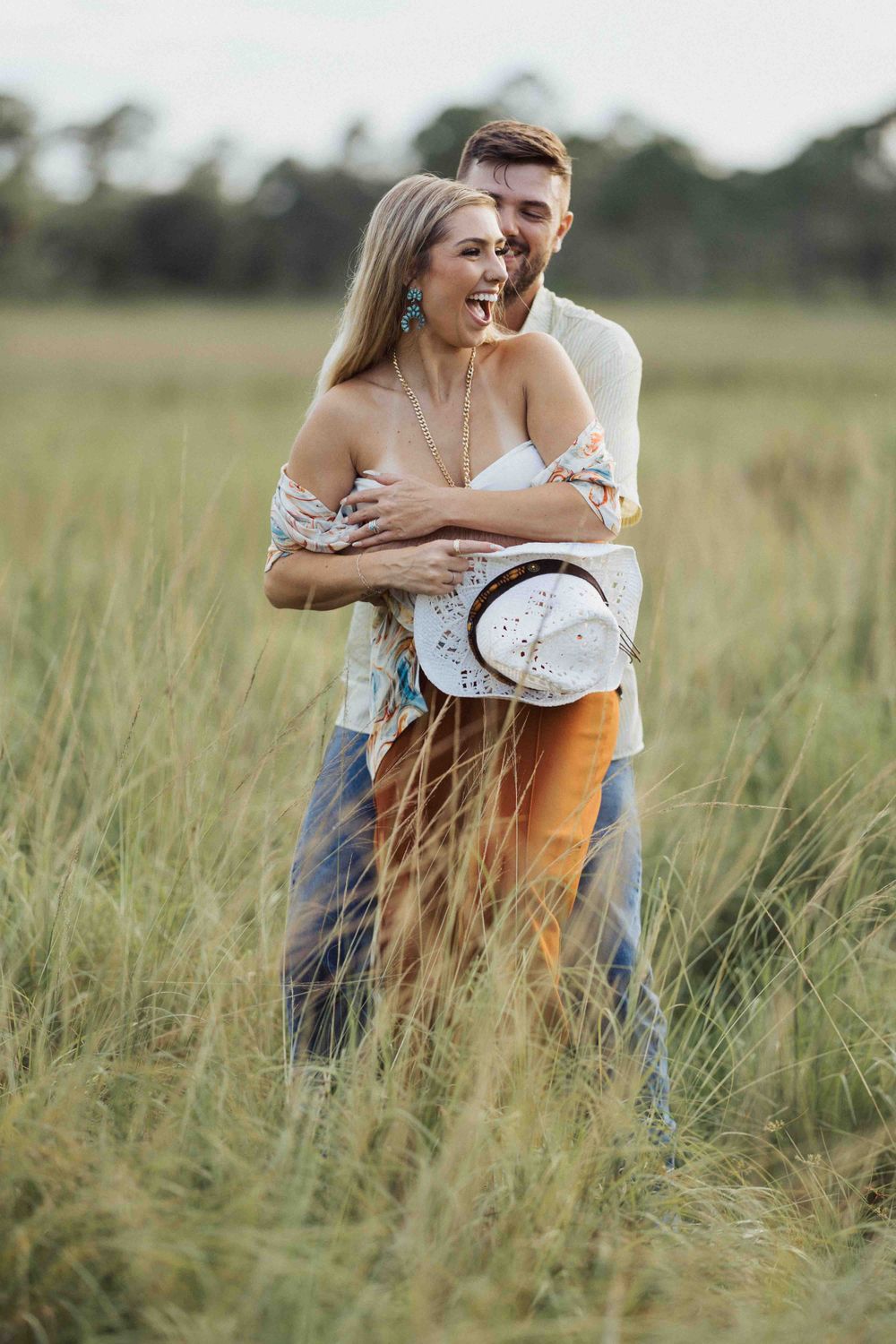 A tender embrace amidst tall grass with a white hat accessory in the golden hour light.