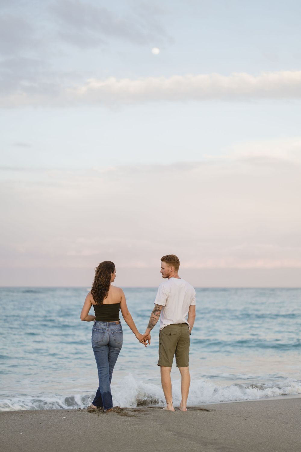 A couple holds hands while standing on a beach at sunset with ocean waves in the background.