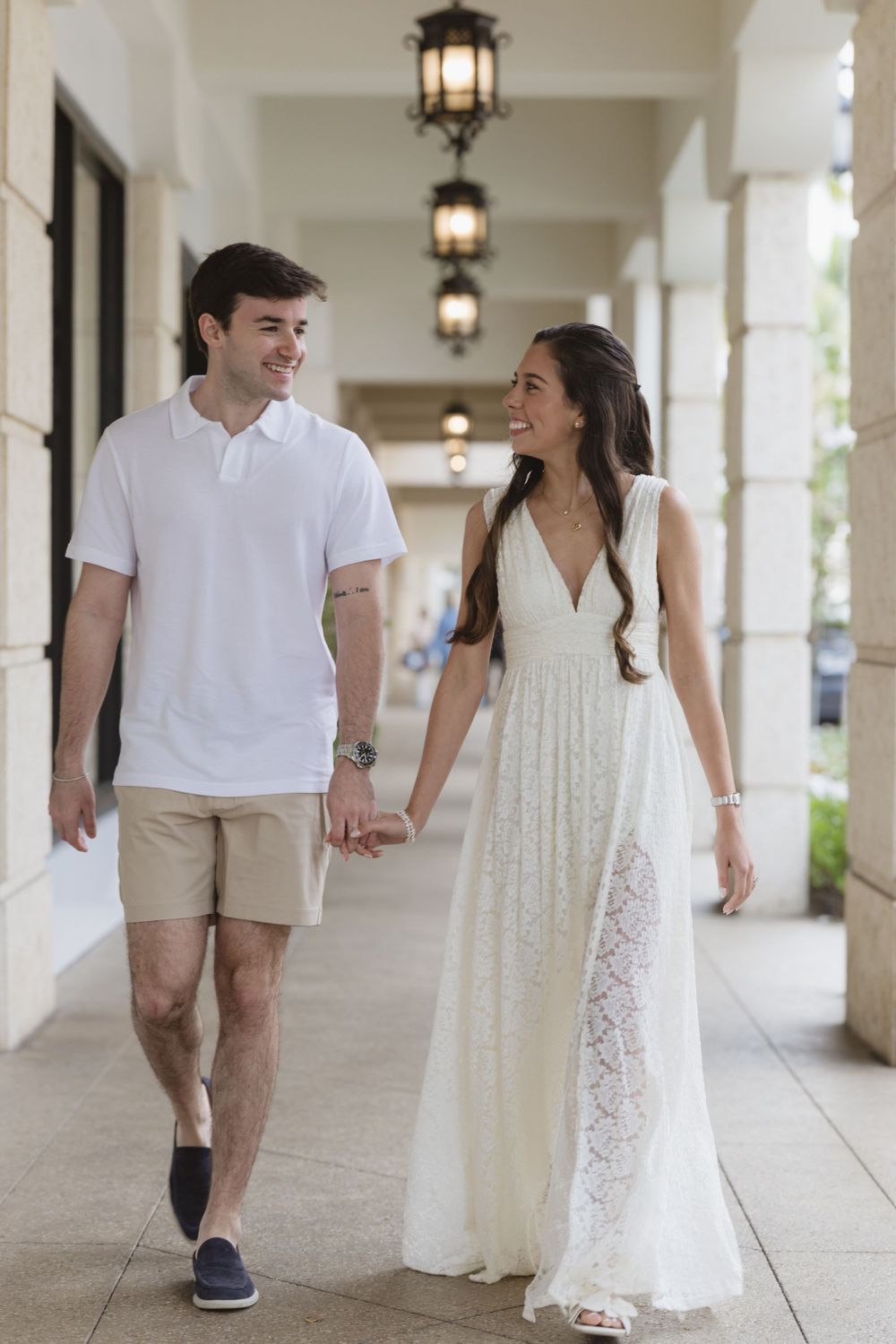A couple walks hand in hand through an elegant colonnade hallway with hanging lanterns.