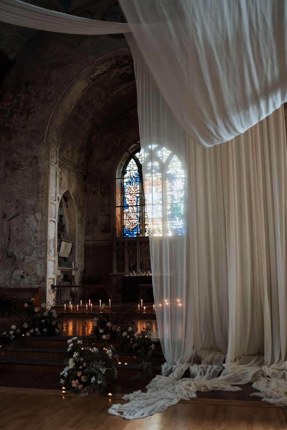 Dramatic stained glass window illuminates a gothic church interior with flowing white drapes and glowing candles.