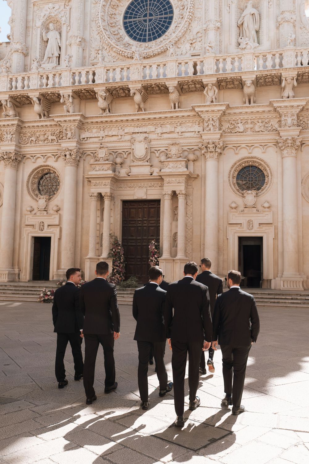 A group in formal black attire walks through an ornate Italian baroque cathedral courtyard with round windows and columns.