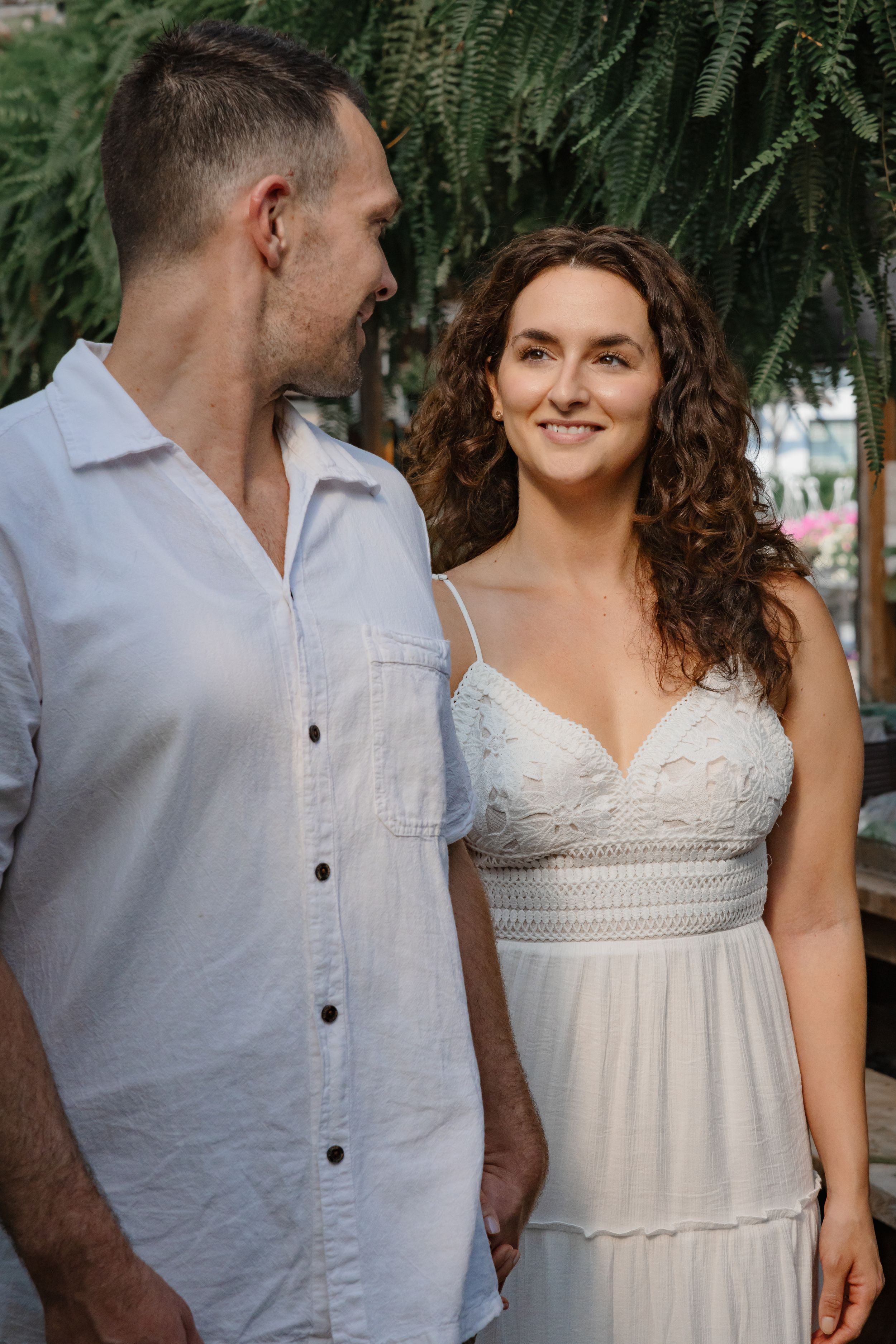 A couple poses together against ferns in casual white attire.