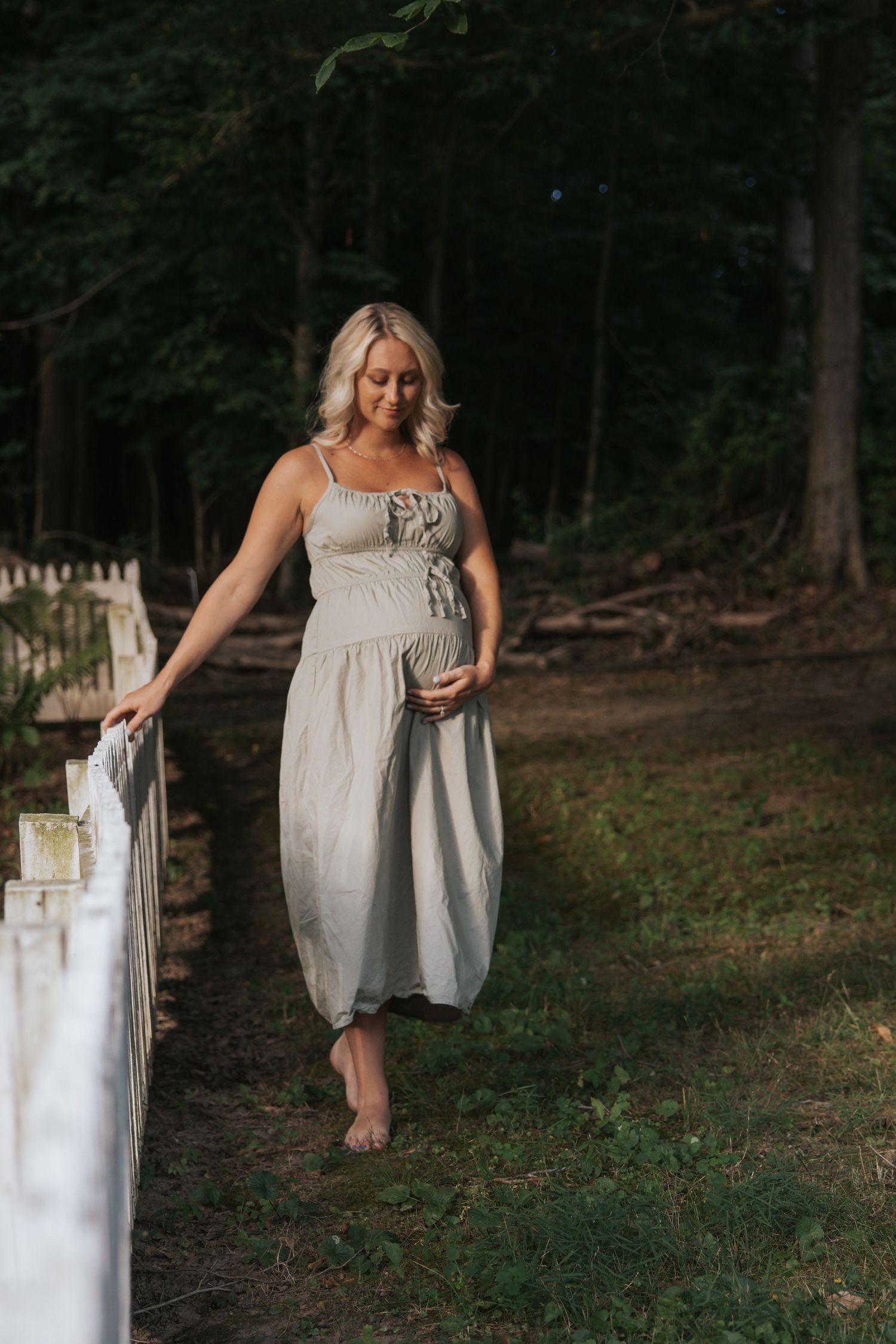 Expectant mother in white dress walks along rustic fence in wooded setting.