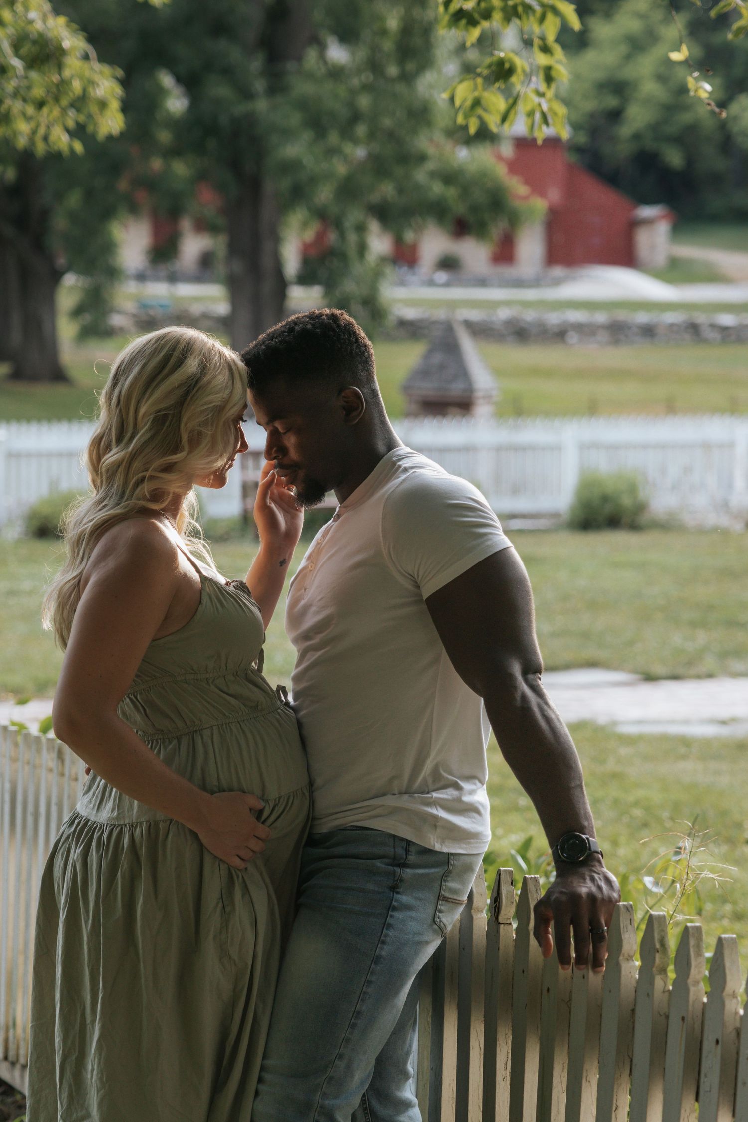 A romantic maternity photoshoot outdoors with a couple standing by a white picket fence and red barn in background.