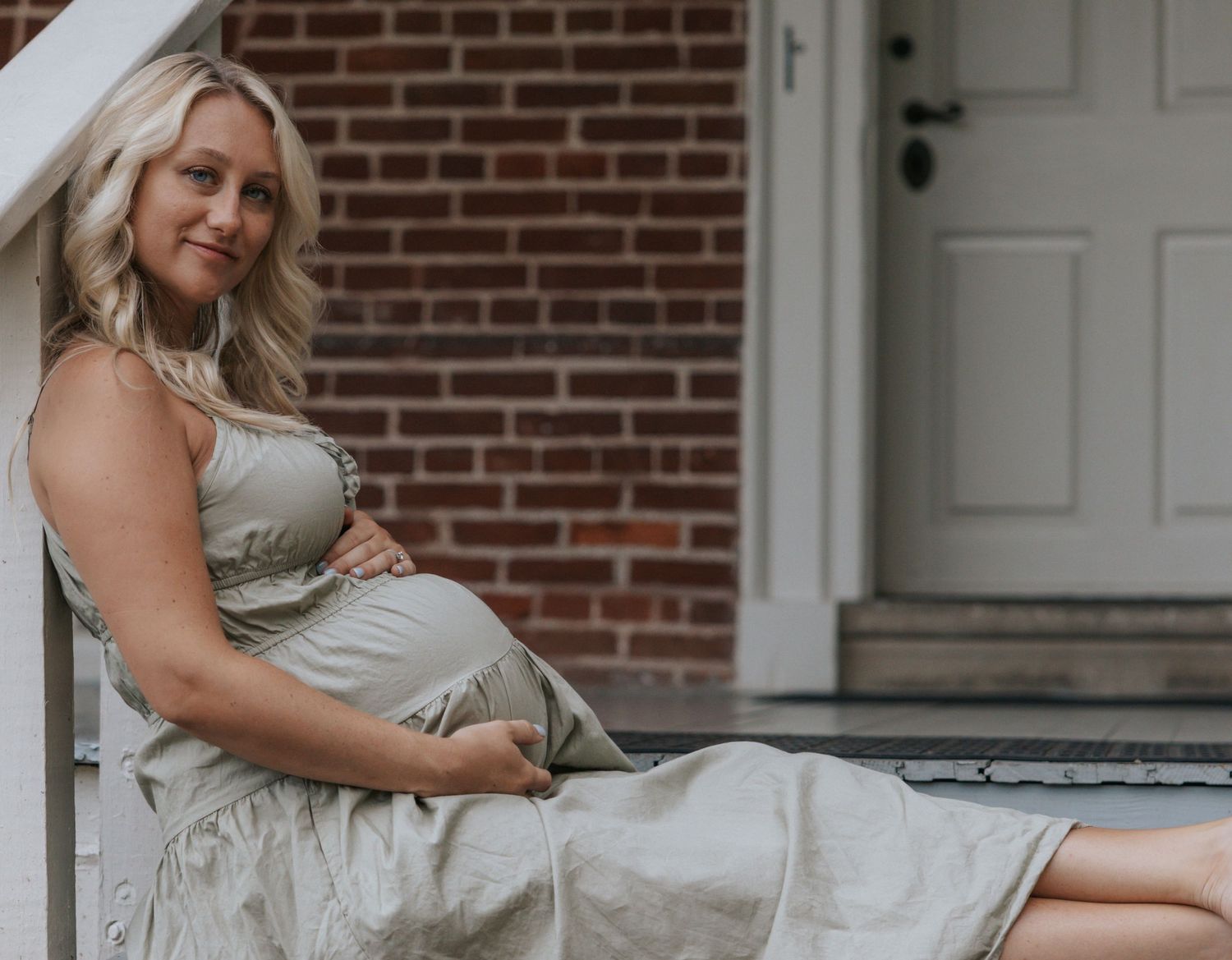 Expecting mother in a sage green dress poses elegantly by a brick wall during maternity photoshoot.