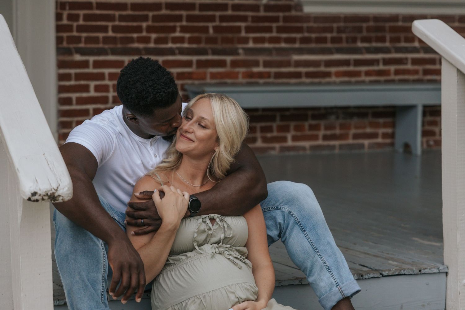 Intimate maternity photo session on a porch with couple sharing tender moment against brick wall.