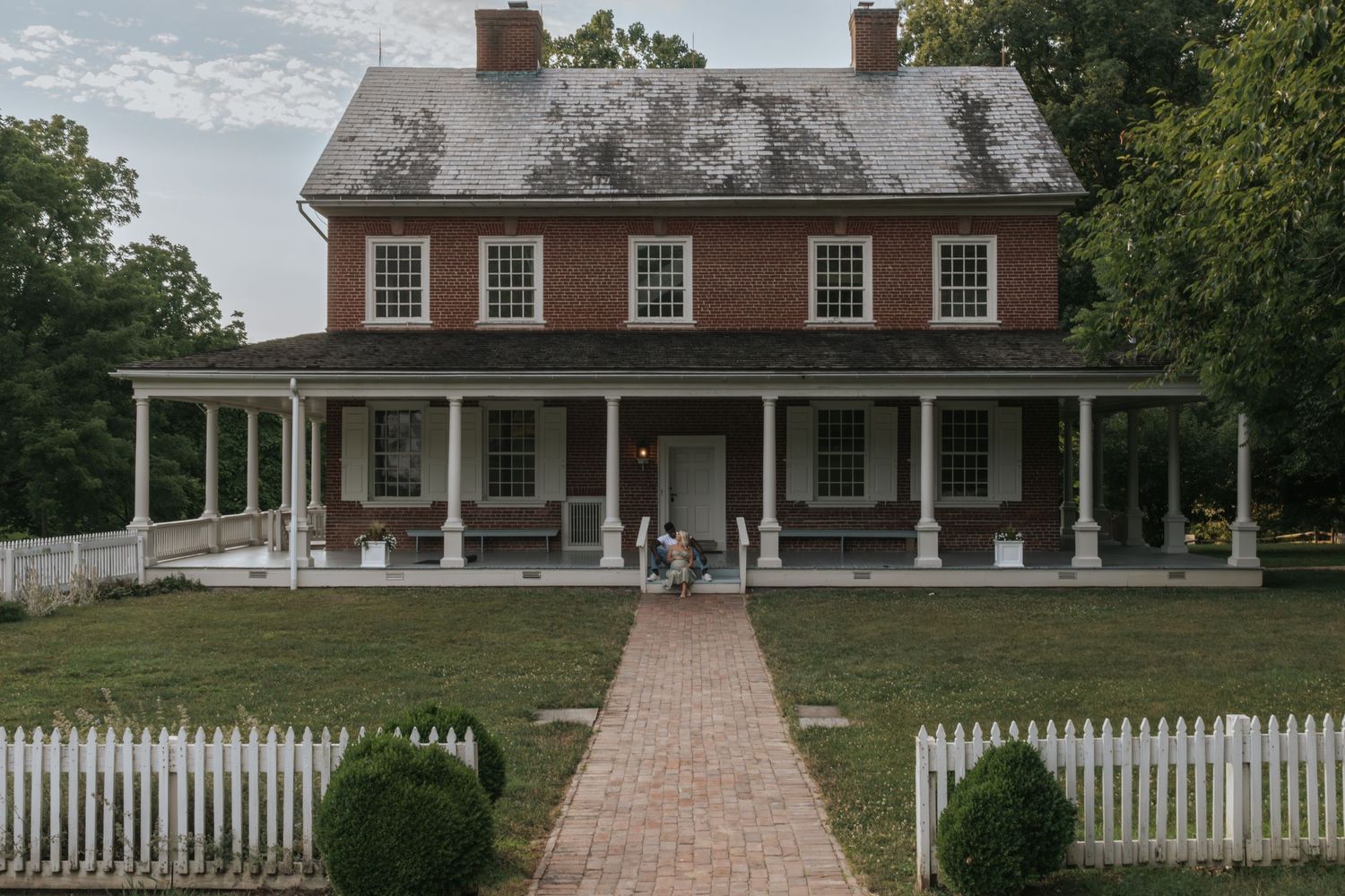 Historic colonial house with white picket fence and brick pathway leading to covered front porch.