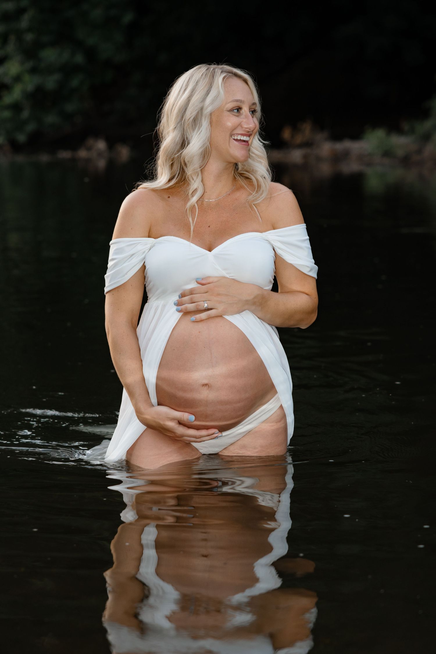 Maternity water photoshoot with woman in white dress standing in lake at sunset.