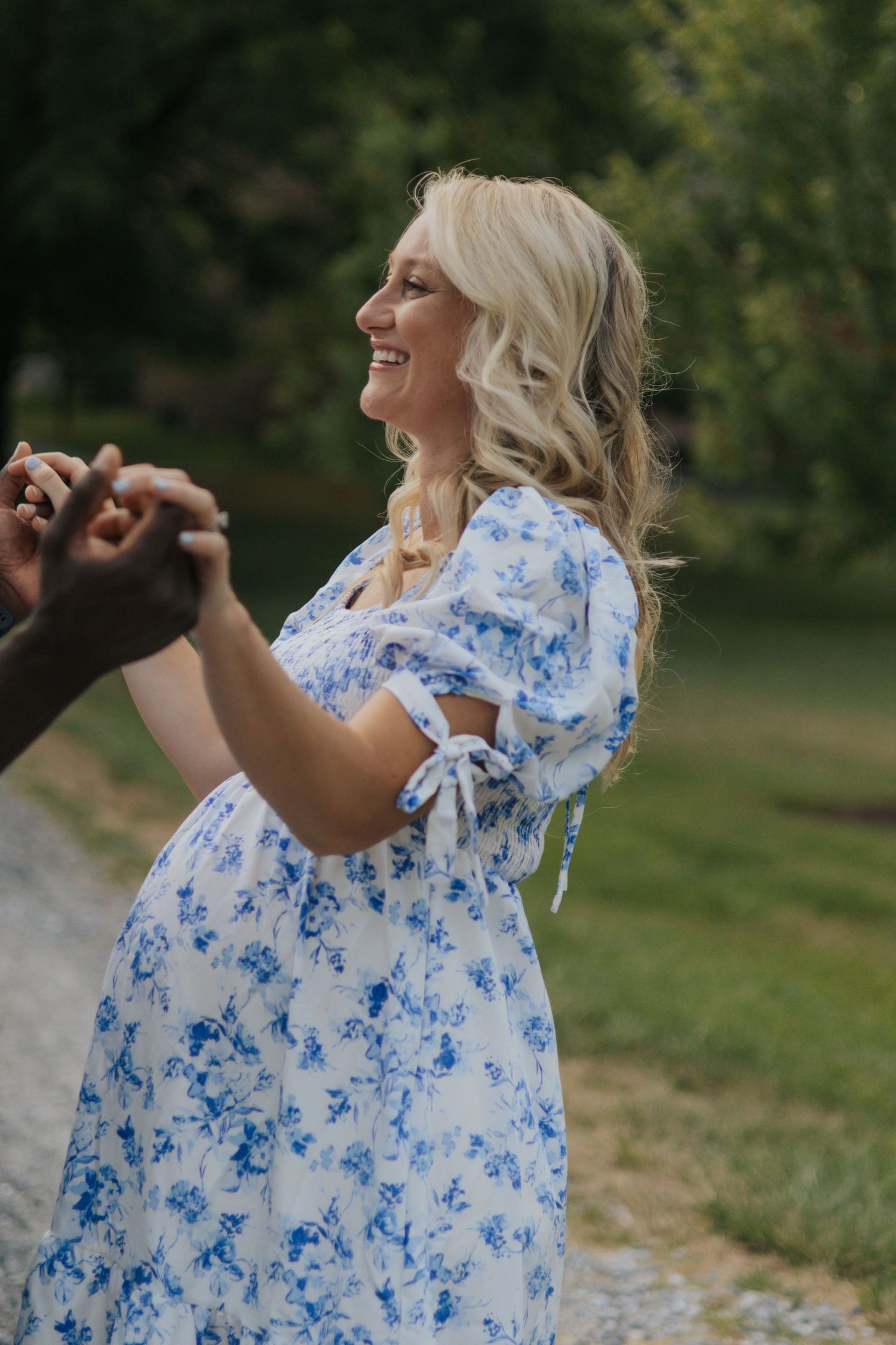 A pregnant woman in a blue floral dress dances joyfully outdoors.