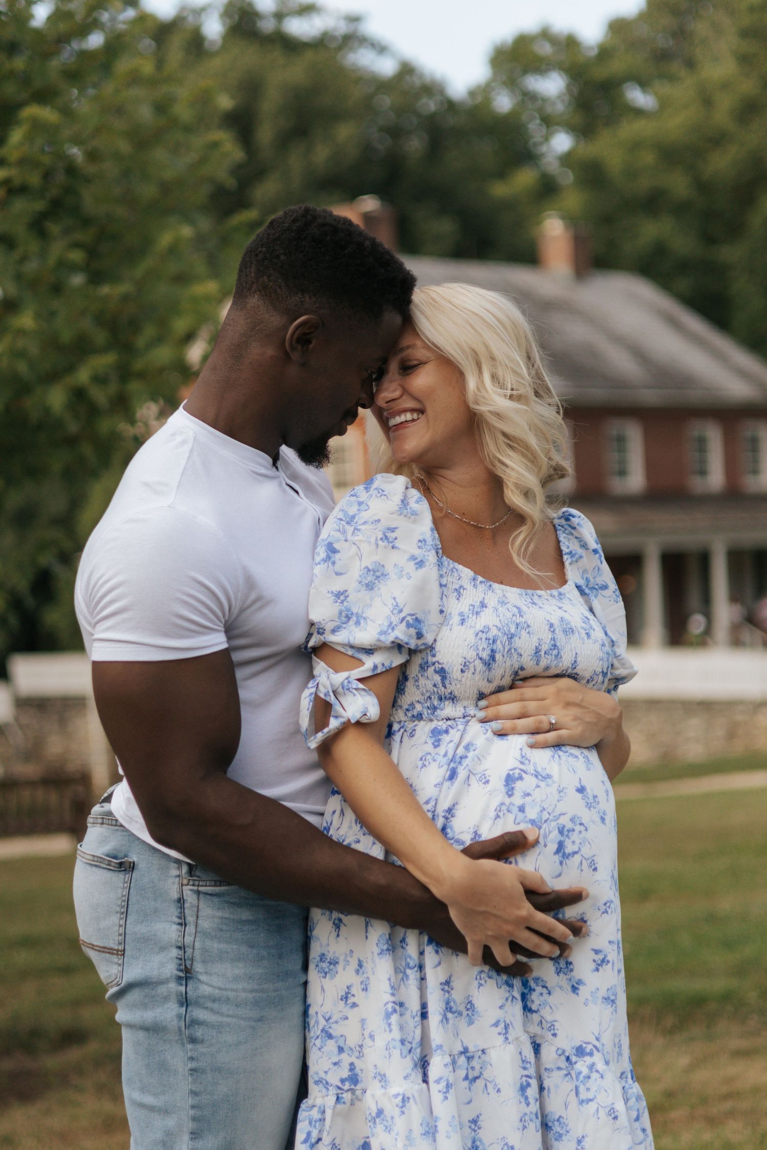 Intimate maternity photo of couple embracing outdoors near a brick house.