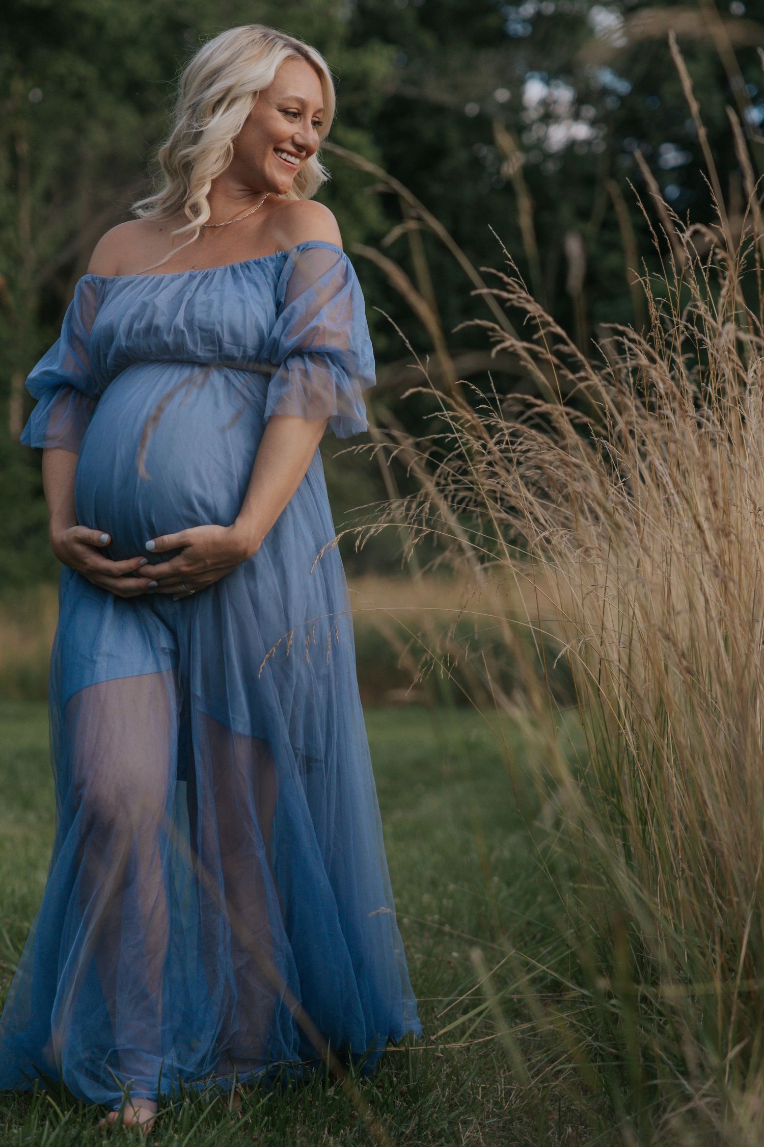 Maternity photo of woman in flowing blue dress standing in tall grass at sunset.