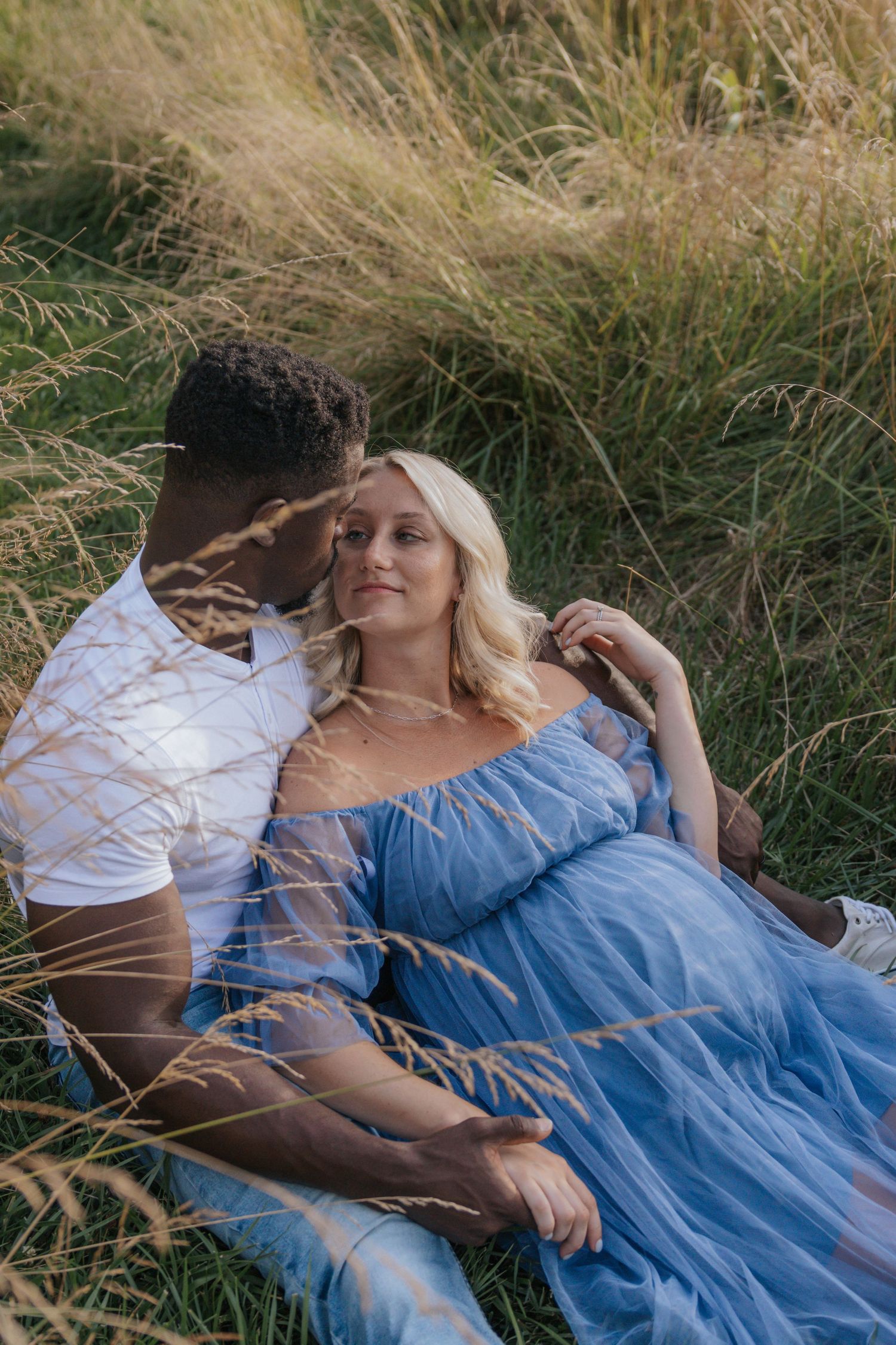 Romantic maternity photo of couple lying in tall grass wearing blue and white.
