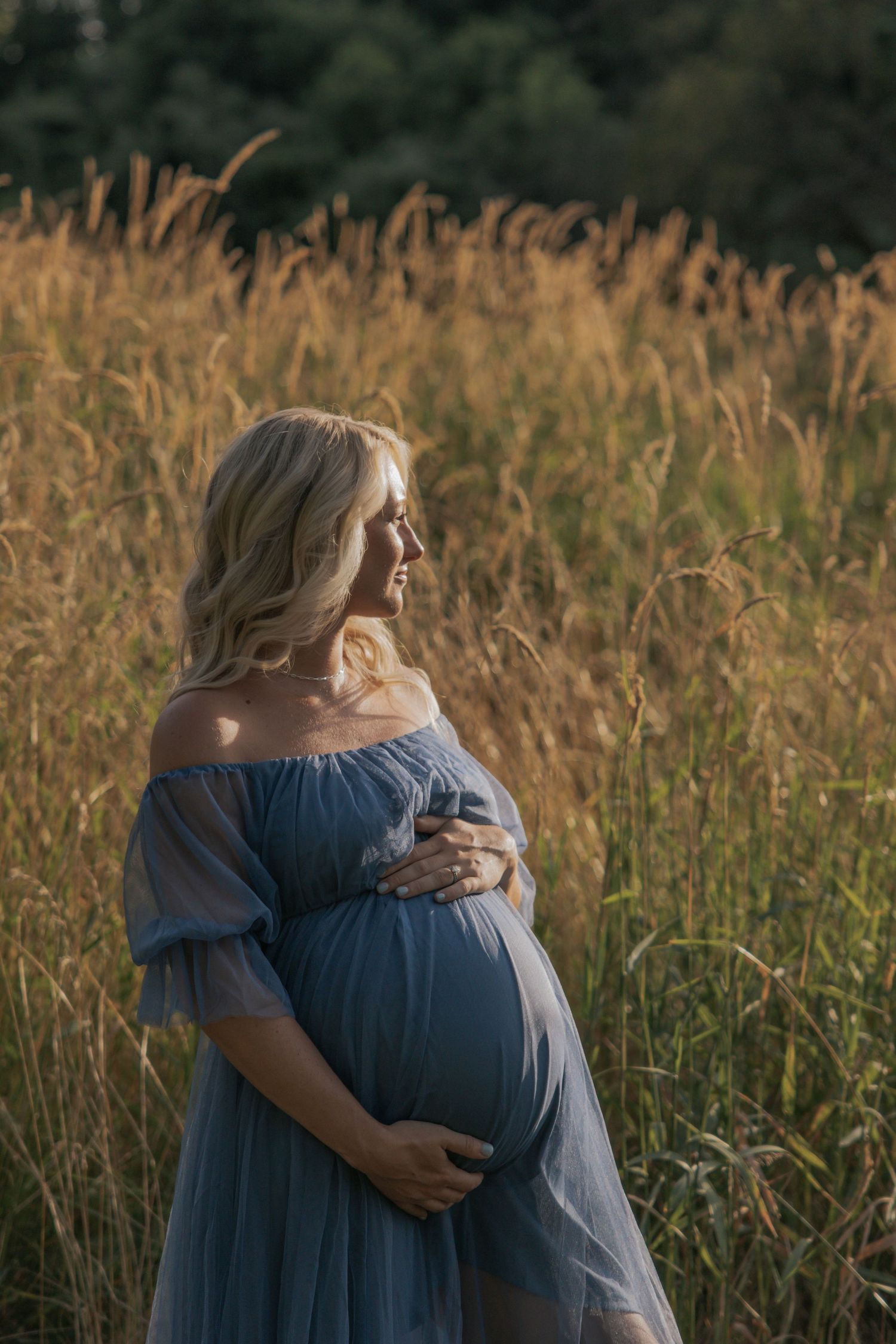 Expectant mother in blue off-shoulder dress poses in golden field at sunset.